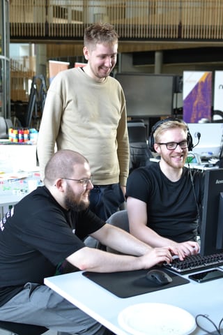 Tríggir luttakarar við eitt arbeiðsborð: ein situr við knappaborðinum við hoyritelefonum, og hinir báðir hyggja við og smíla.
